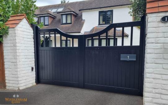 Black wooden electric driveway gates with a distinctive asymmetric arched top and vertical panelling, featuring an open slat design in the upper section and integrated letterbox, set between white-painted brick pillars with tiled caps; by Windlesham Gates