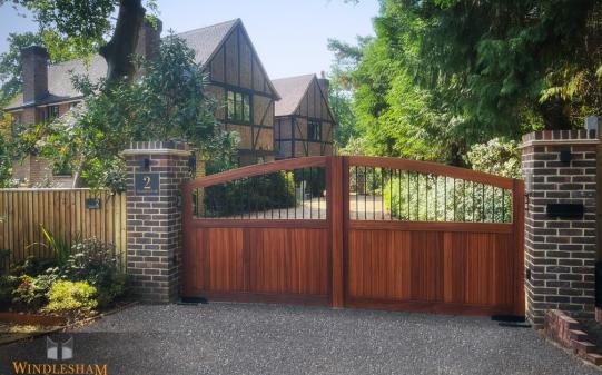 Timber electric driveway gates with a warm wood finish, arched top, and vertical slat lower section paired with open metal infills above, set between dark brick pillars at the entrance to a characterful Tudor-style home; by Windlesham Gates