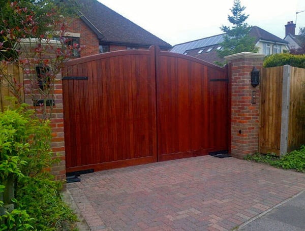 Solid wooden electric driveway gates with a gentle arched top and vertical panel finish, mounted between red brick pillars, leading to a residential home in the UK.