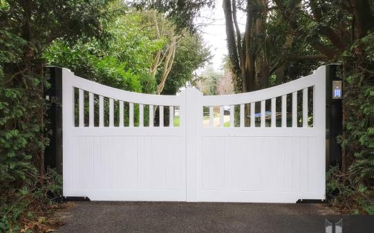 White-painted wooden electric driveway gates with a gentle inward curve and vertical slat top section, set between dark posts and surrounded by mature greenery, offering a stylish and private entrance to a secluded UK property.