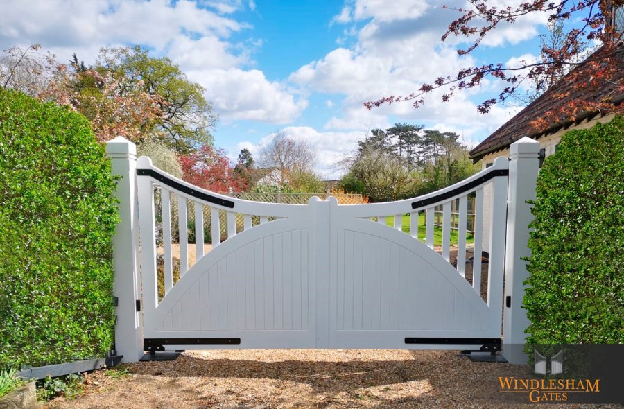 White wooden electric driveway gates with a sweeping upward curve and arched lower panel design, featuring vertical slats and black hinge detailing, set between square posts and surrounded by neatly trimmed hedges at the entrance to a charming rural UK property; by Windlesham Gates