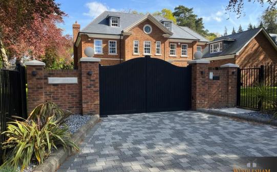 Black wooden electric driveway gates with a symmetrical double-arched top and vertical slat design, set between red brick pillars topped with stone ball finials and flanked by wrought iron fencing, providing a secure and elegant entrance to a large Georgian-style UK home; by Windlesham Gates