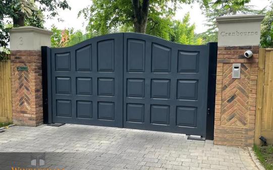 Dark grey wooden electric driveway gates with an elegant arched top and raised rectangular panel design, set between decorative brick and rendered pillars at the entrance to a home named "Cranbourne"; by Windlesham Gates