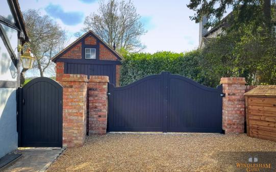 Black wooden electric driveway gates with a flowing double-curved top and vertical slat design, accompanied by a matching pedestrian gate, both set between rustic red brick pillars and enclosing a gravel driveway of a charming UK cottage-style property; by Windlesham Gates