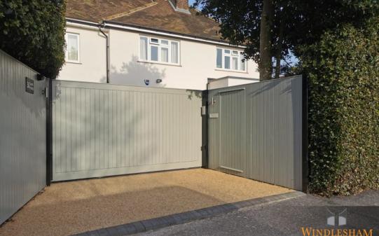 Light grey aluminium bi-fold electric driveway gates with a clean vertical slat design, offering a space-saving solution for narrow entrances, installed at the front of a modern UK home with gravel driveway; by Windlesham Gates
