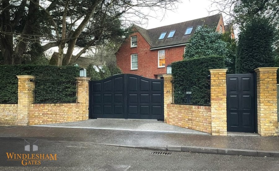 Elegant black wooden electric driveway gates with a raised arched centre and classic square panel design, paired with a matching pedestrian gate, both set between yellow brick pillars with stone caps and black wrought iron accents, securing a red brick period home; by Windlesham Gates