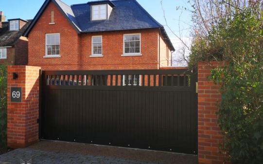 Modern black aluminium sliding electric gate with vertical panelling and an open slat top section, installed between red brick pillars at the entrance to a traditional-style red brick home with a slate roof, marked with house number 69.