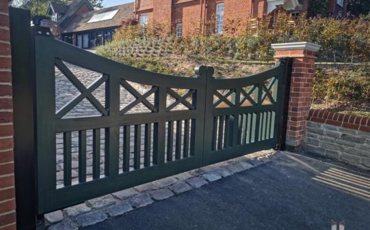 Dark green wooden electric driveway gates with a sweeping curved top, featuring decorative cross-brace and vertical slat detailing, set between red brick pillars at the entrance to a cobbled drive leading to a traditional UK property.