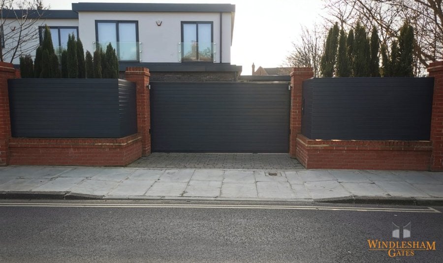 Modern black aluminium sliding electric gate with sleek horizontal lines, seamlessly integrated with matching privacy panels and red brick pillars, providing secure and stylish access to a contemporary UK home; by Windlesham Gates