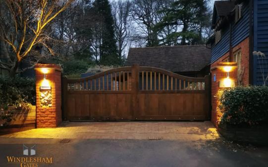 Warm-toned wooden electric driveway gates with a gentle arched top and vertical slat design, beautifully lit by wall-mounted lanterns on rustic brick pillars, creating a welcoming and secure entrance to a countryside property at dusk; by Windlesham Gates