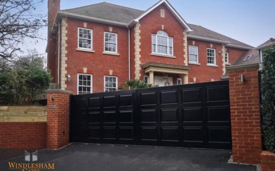 Tall black wooden electric driveway gates with a flat top and classic raised square panel design, perfectly complementing the grand red brick Georgian-style home behind, and mounted between red brick pillars; by Windlesham Gates