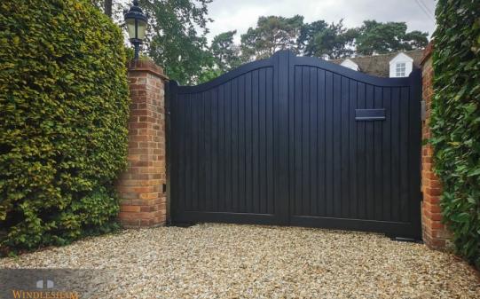 Black wooden electric driveway gates with a graceful double-curved top and vertical slat design, featuring an integrated letterbox, set between traditional red brick pillars with lantern lights, leading to a gravel driveway of a classic UK property; by Windlesham Gates